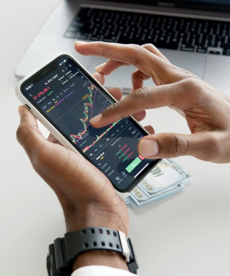 Hands using smartphone trading app with charts, dollar bills in background.